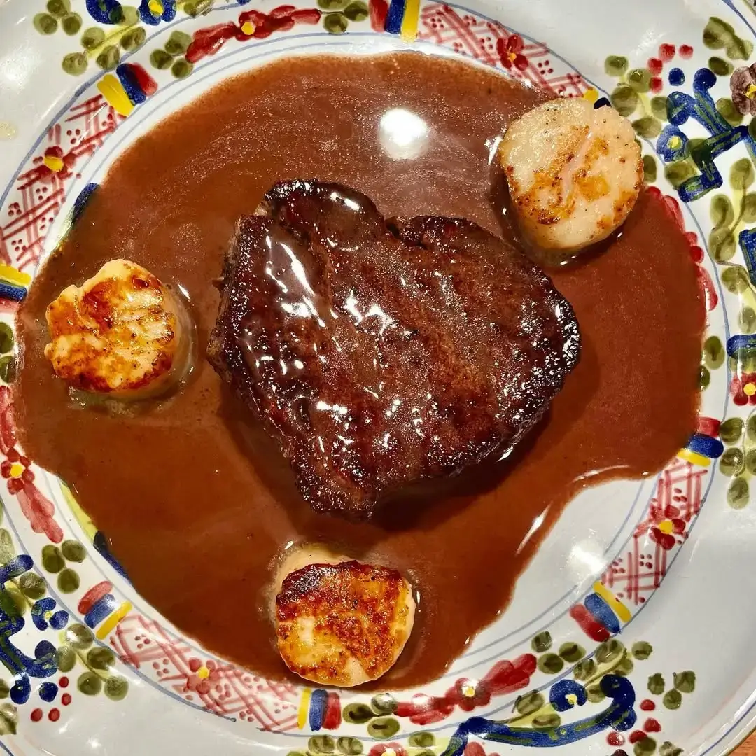 A sauced steak course plated with seared scallops on a painted ceramic plate.
